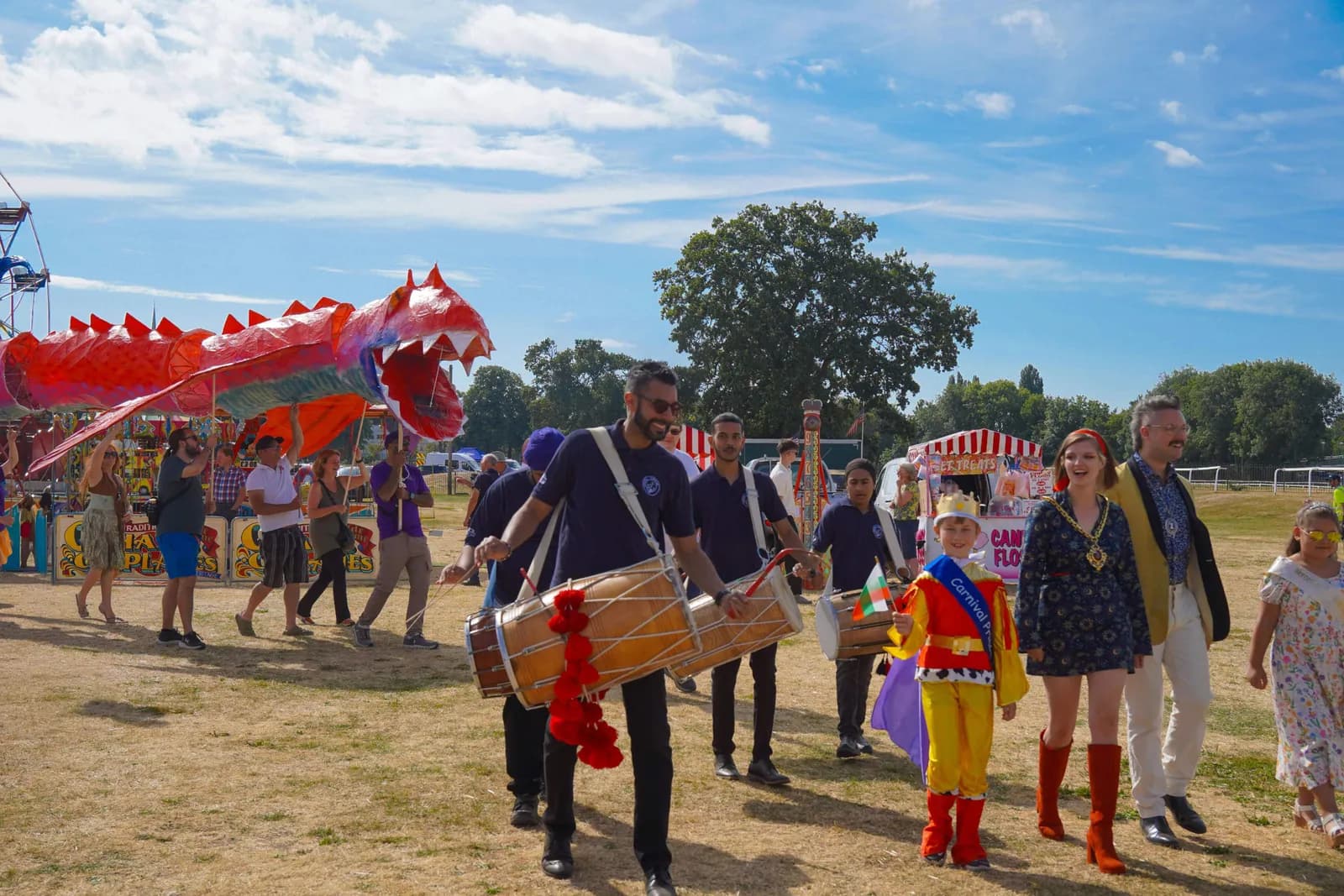A parade with people dressed in costumes, including a large red dragon puppet, walking on a grassy field under a partly cloudy sky.