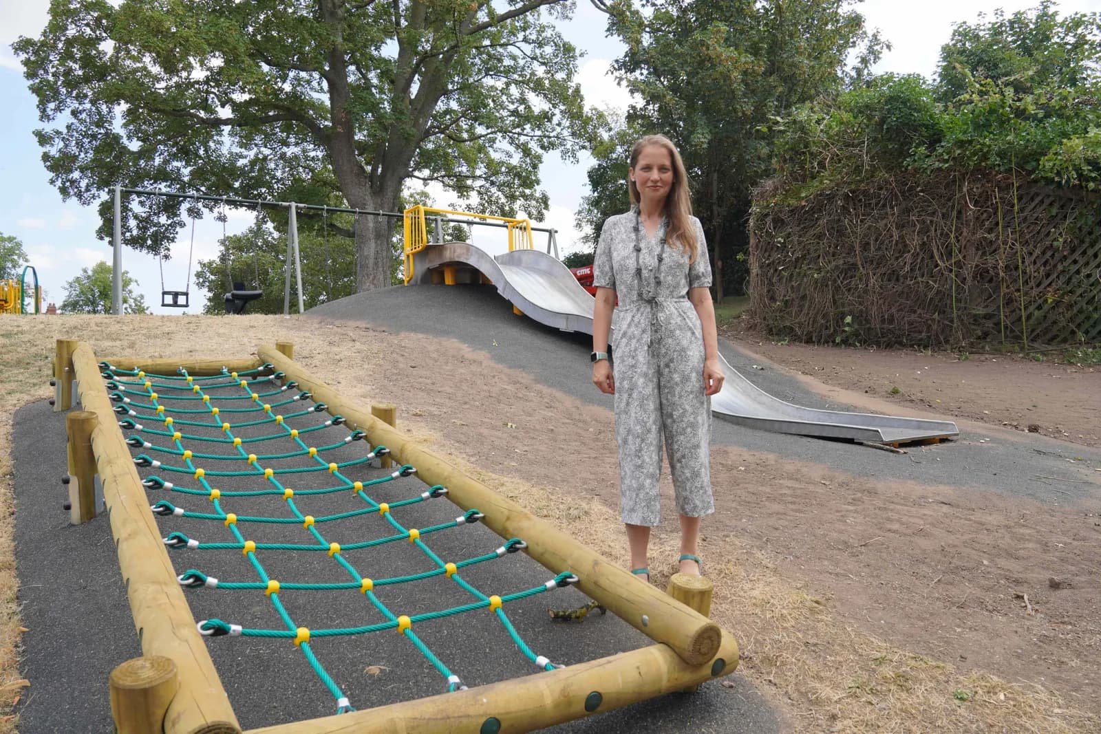 A woman stands in a gray jumpsuit on a gravel slope, beside a climbing net and slide in a playground with trees in the background.