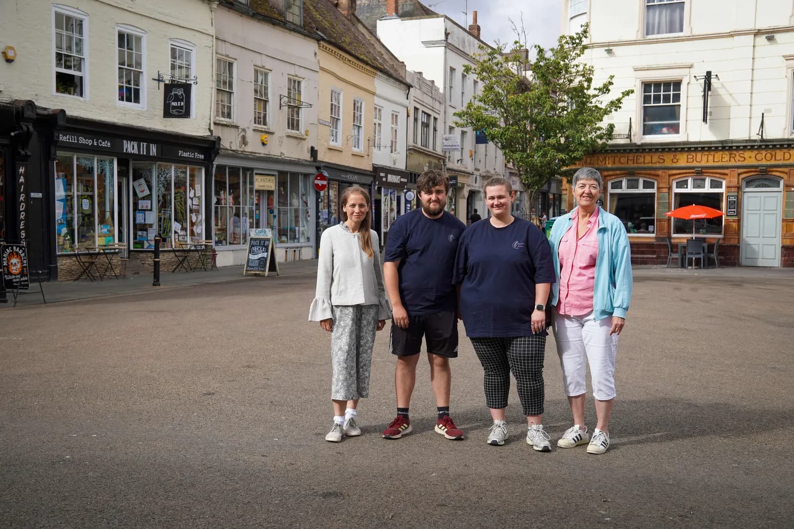 Four people, two men and two women, stand in a row facing the camera on a paved street in front of a row of shops and buildings.