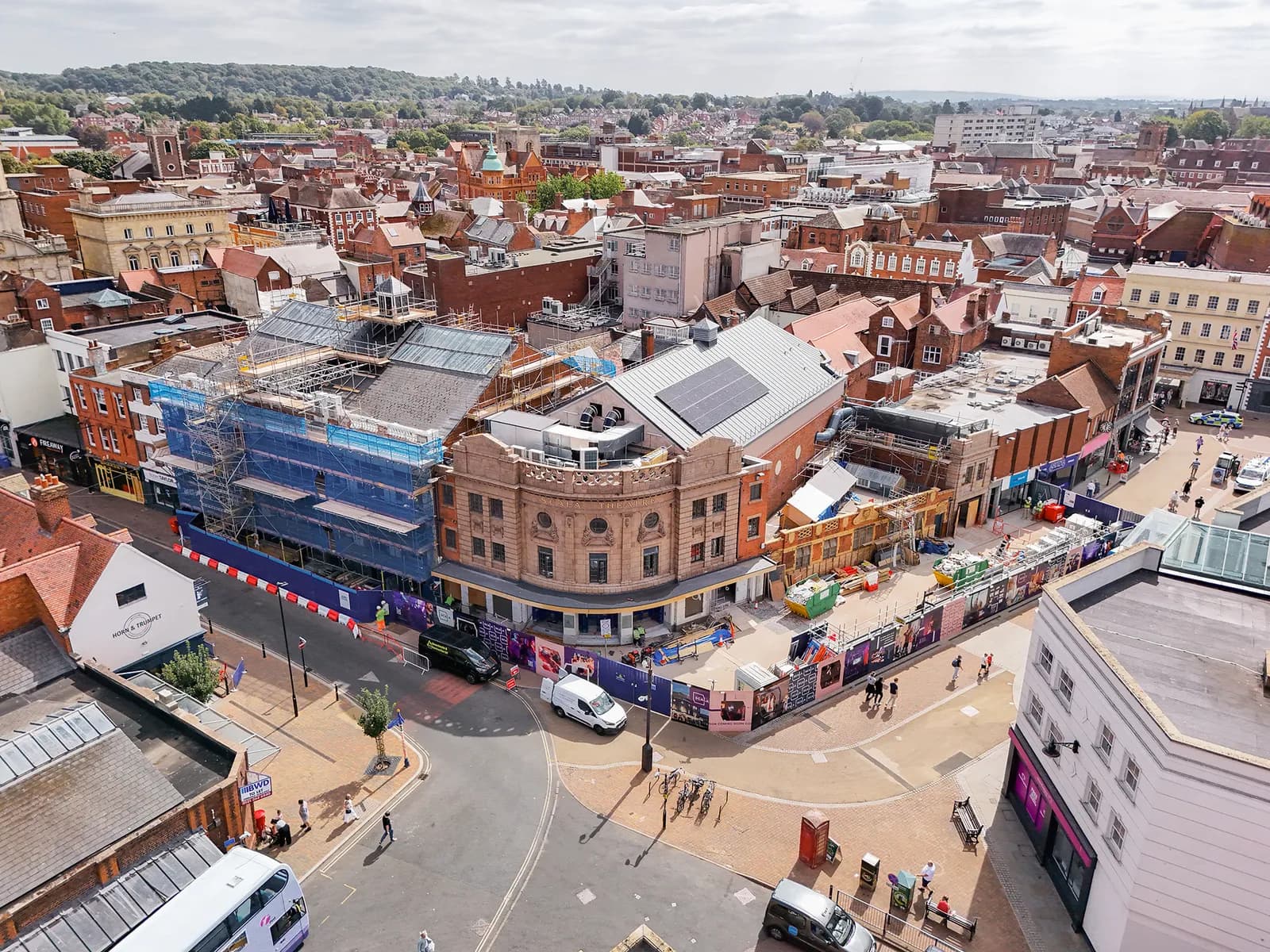 An aerial view of a busy town center with many buildings, including one covered in blue scaffolding, and streets with cars and pedestrians.