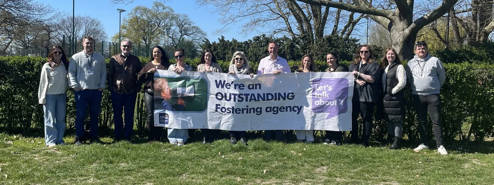 A group of thirteen adults stands in a grassy area holding a banner that reads "We're an OUTSTANDING Fostering agency. Let's talk about it."