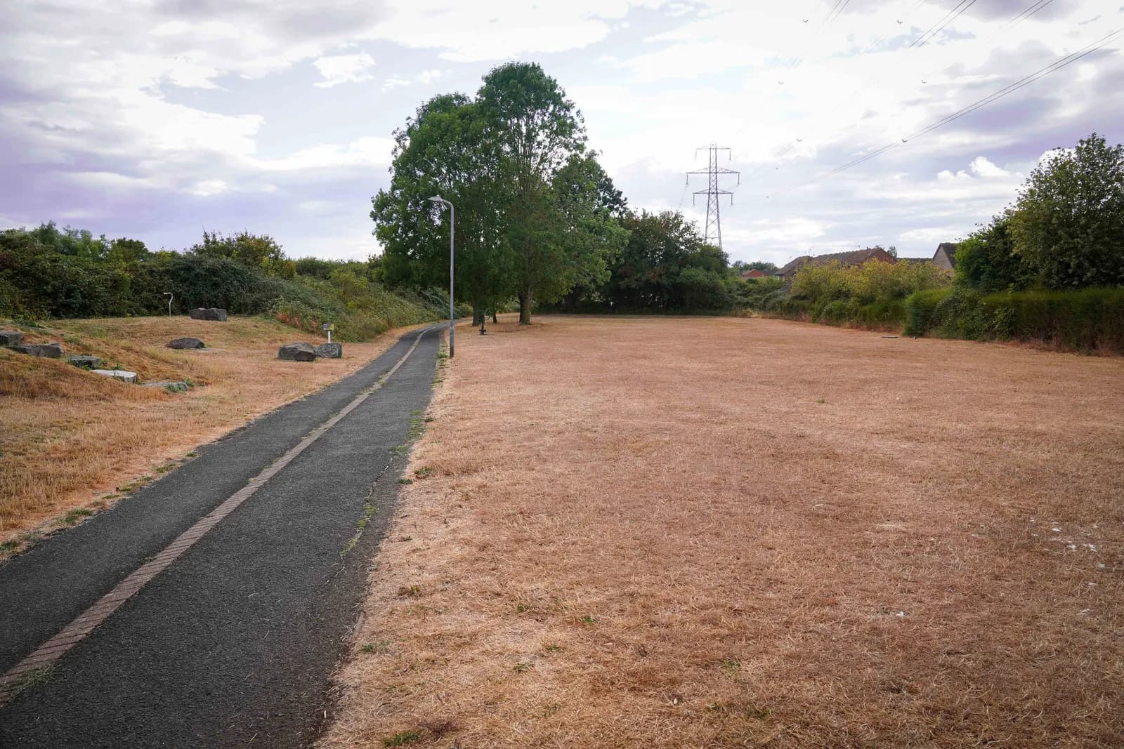 A long, narrow asphalt path runs alongside a hill covered in dry, brown grass and green bushes, leading to a large field of parched grass under a cloudy sky.