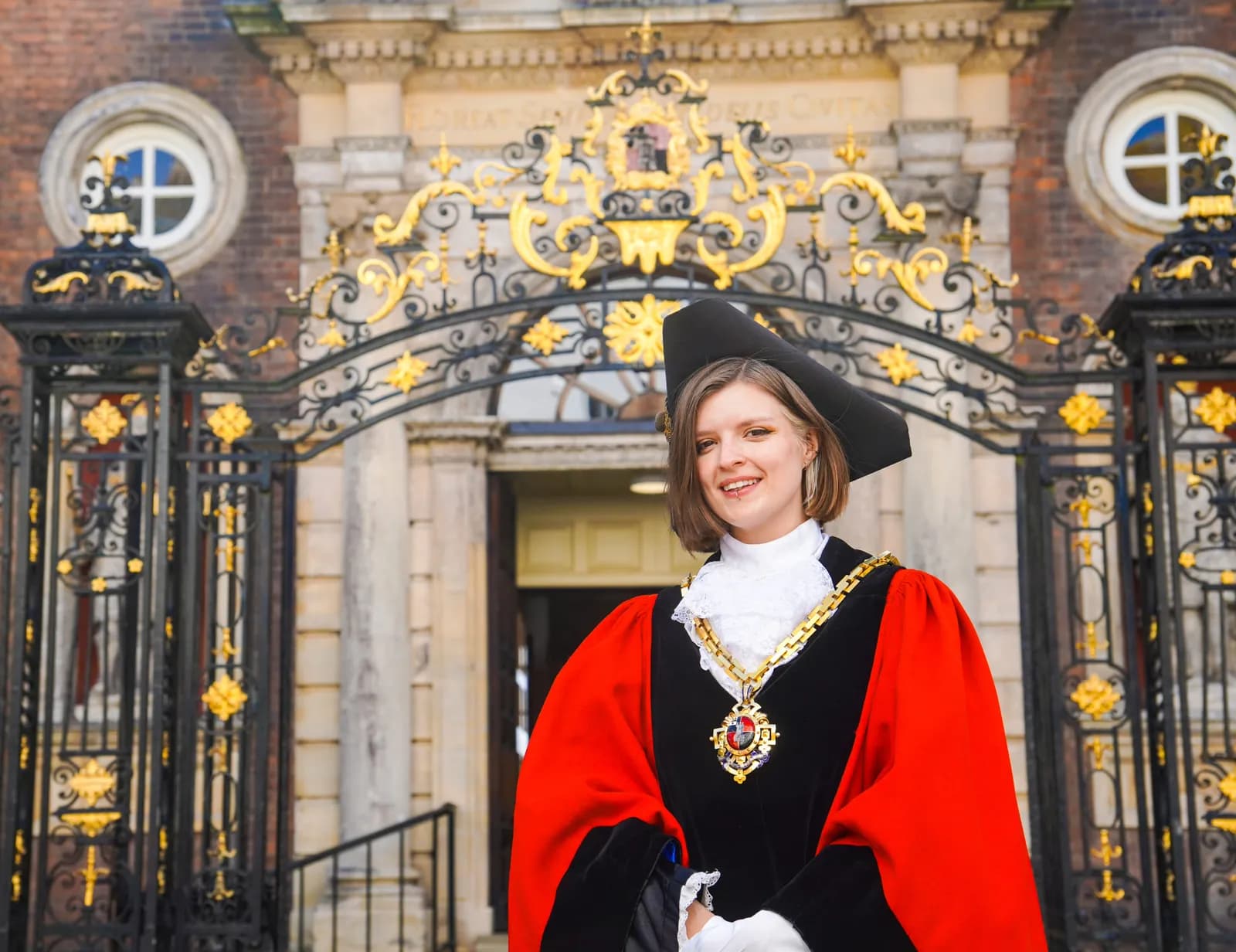 A young woman in traditional academic or civic ceremonial robes and a hat stands smiling in front of ornate iron gates and a stone building.