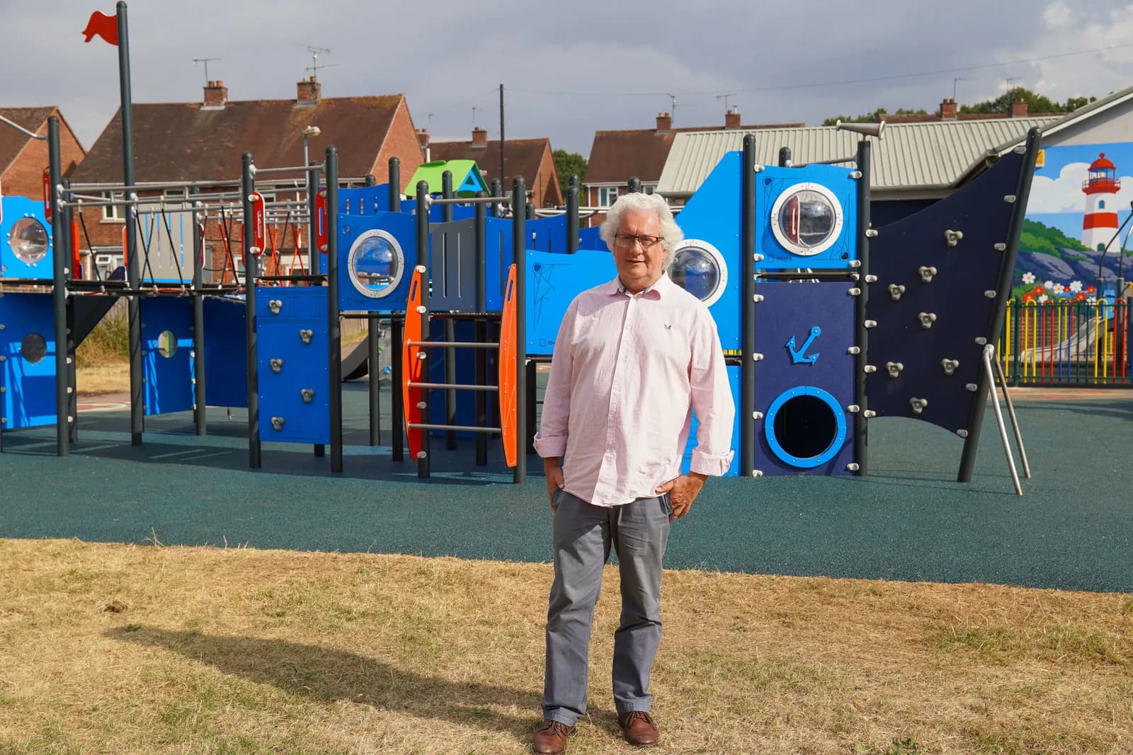 A man with white hair wearing a pink shirt and grey pants stands in front of a blue and orange playground structure.