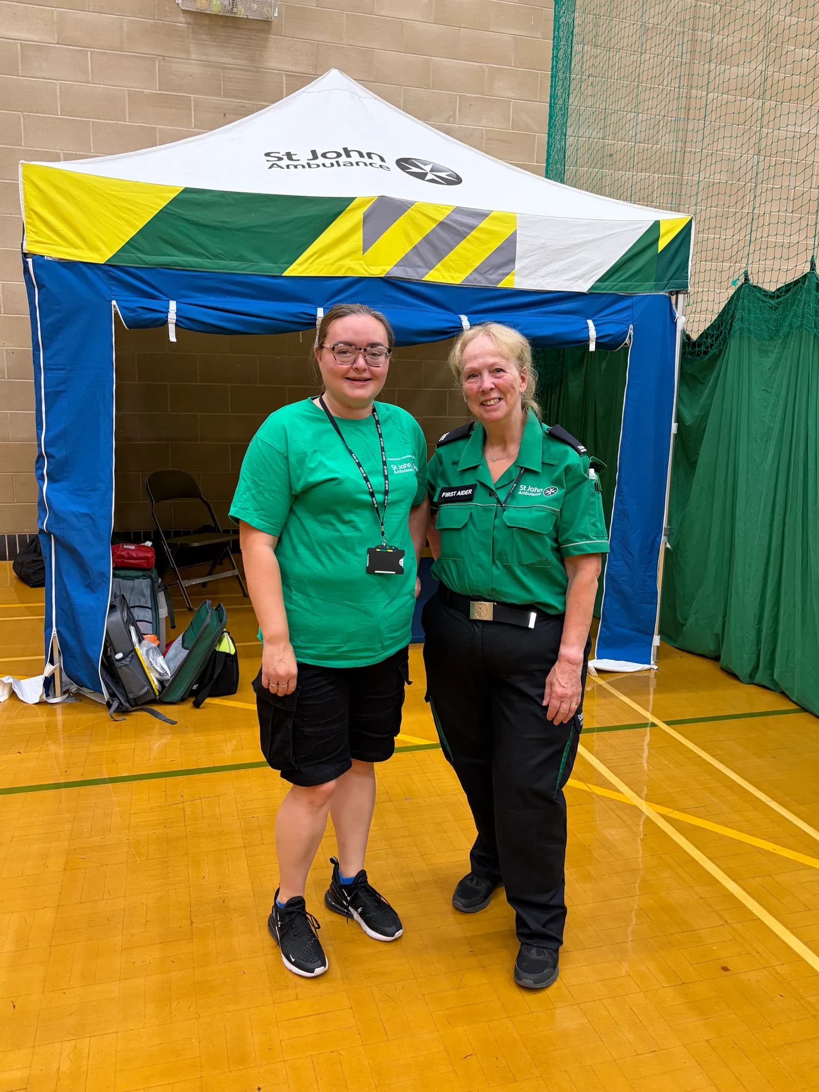 Two women, one with dark hair and glasses and another with blonde hair, both wearing St John Ambulance uniforms, stand smiling in front of a white and green St John Ambulance tent.